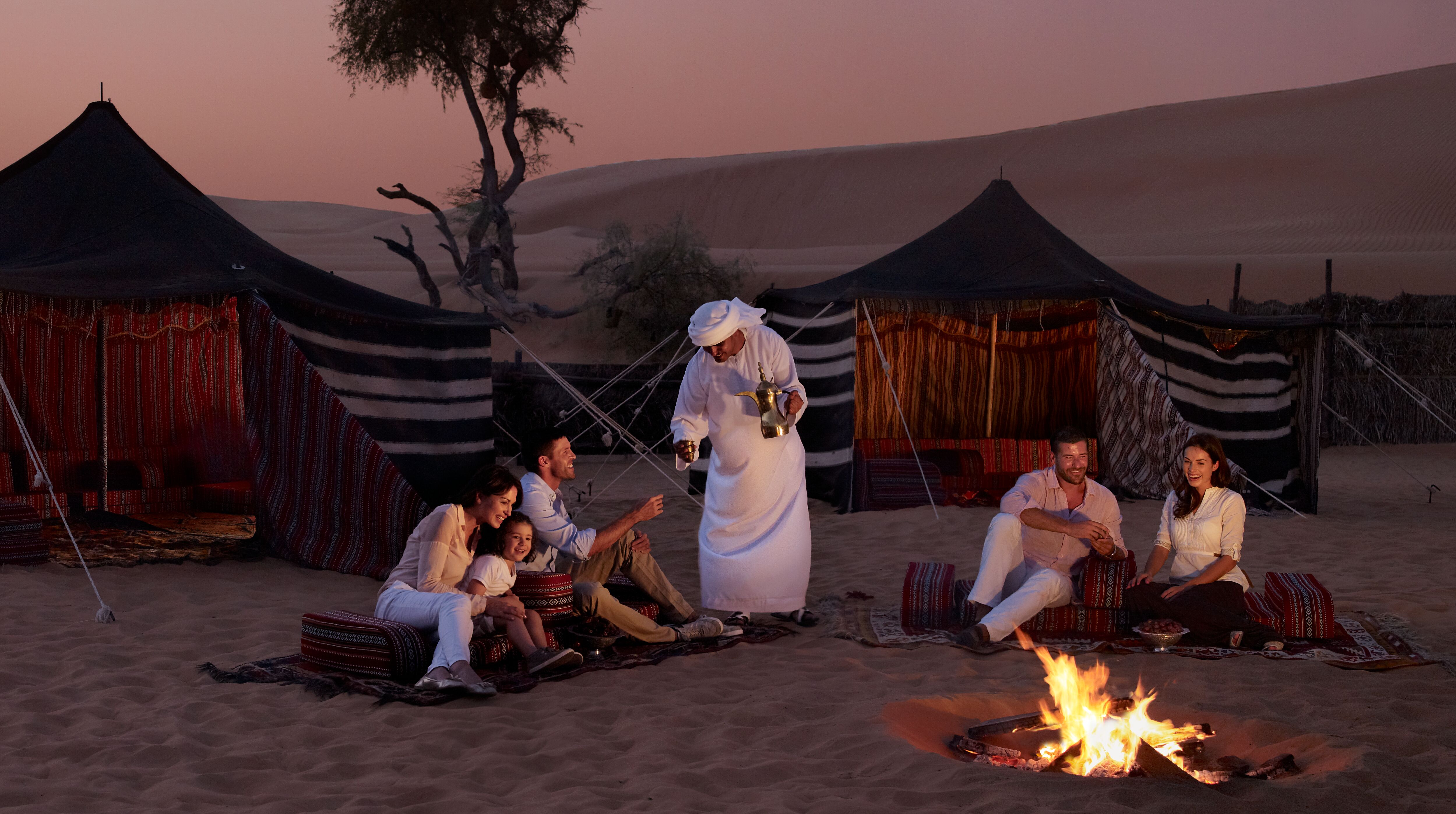 كثبان رملية أمامها بيوت شعر وأشخاص أجانب يحتسون القهوة المقدمة لهم من قبل رجل بزي إماراتي حول النار المضاءة في  الوسط وقت المغرب