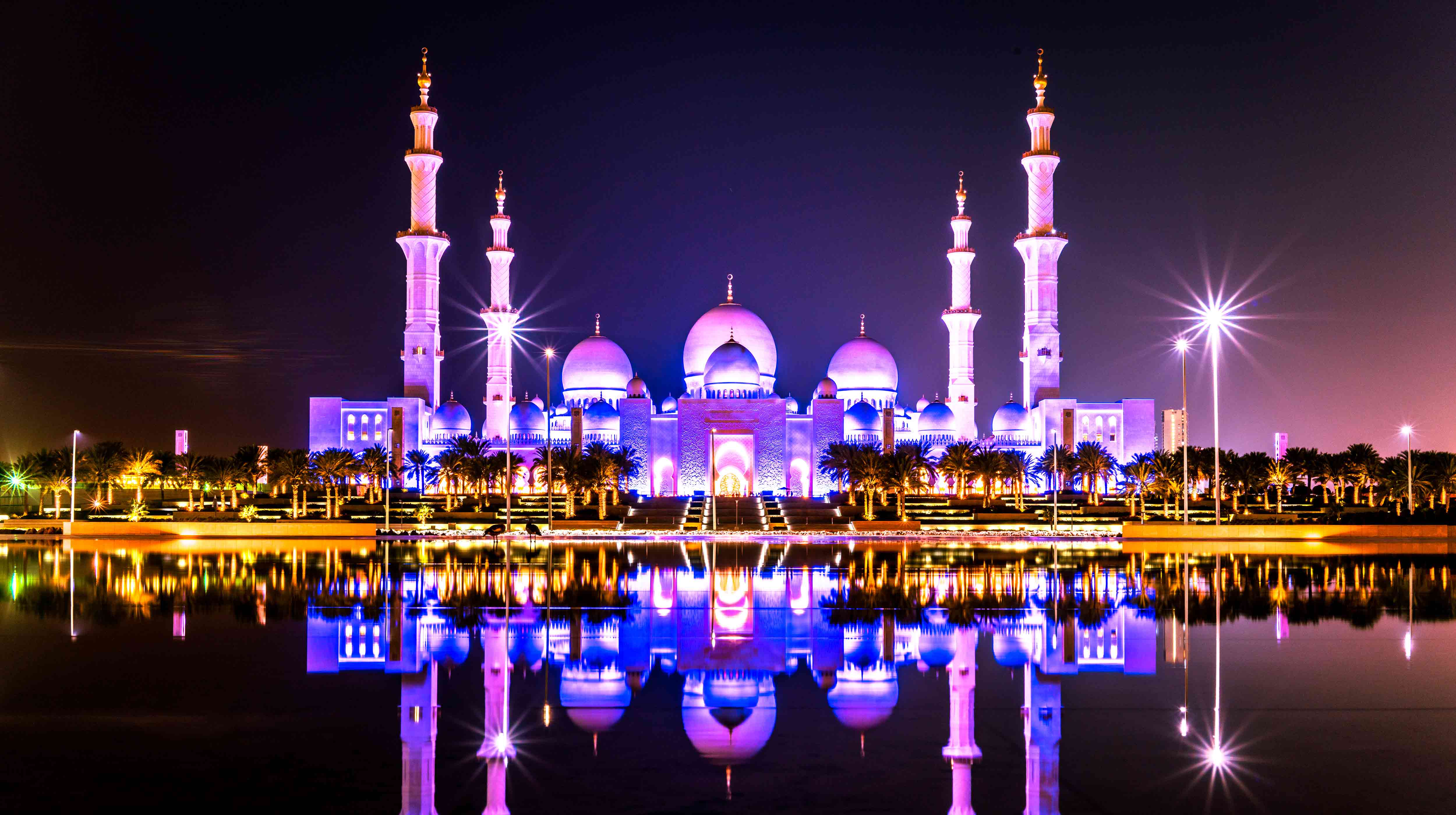 جامع الشيخ زايد الكبير في الليل منار باللون البنفسجي أمامه أشجار النخيل والبحيرة ومضاء بالأنوار 